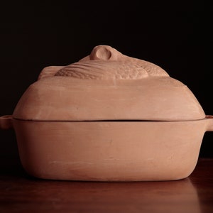 May include: A terracotta casserole dish with a lid. The lid has a decorative design of a bird.