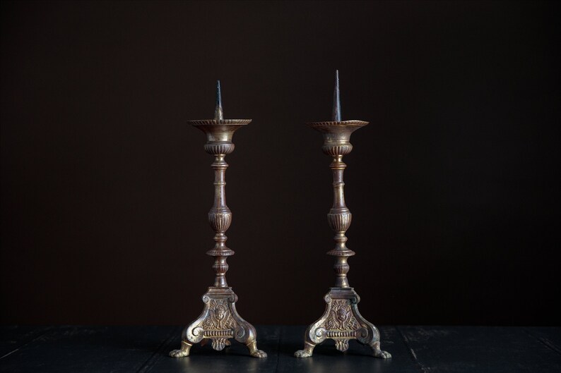 Puede incluir: Un par de candelabros antiguos de lat&oacute;n con detalles ornamentados y un acabado marr&oacute;n oscuro. Los candelabros est&aacute;n sobre una superficie negra.