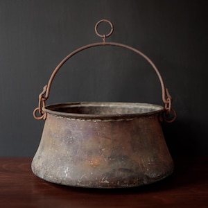 May include: A rustic, antique copper cauldron with a curved handle and a small ring at the top. The cauldron has a wide, rounded body and a weathered, aged appearance. The metal has a dark, mottled patina, suggesting age and use. The cauldron sits on a dark wooden surface.