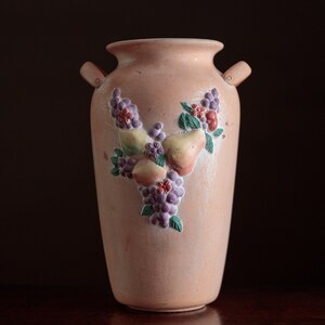 May include: A light pink ceramic vase with a floral design featuring pears, grapes, and flowers. The vase has two handles on the sides.