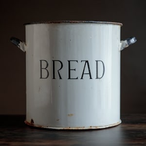 May include: White metal bread bin with black handles and the word "BREAD" printed on the front. The bin has a worn, vintage look.