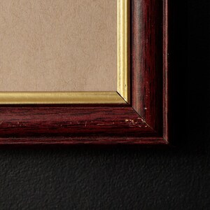 May include: Close-up of a framed artwork. The frame is a dark reddish-brown with a gold inner edge. The artwork itself is a light tan color. The frame has some minor wear and tear. The background is black.
