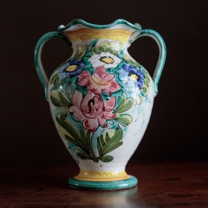 May include: A white ceramic vase with green and yellow accents. The vase has two handles and is decorated with a floral pattern in shades of pink, blue, and green.
