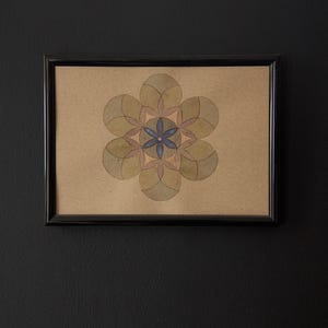 May include: A framed artwork featuring a symmetrical floral design. The artwork is composed of overlapping petal shapes in shades of green and brown, with a blue center. The frame is black, and the background is a dark wall.