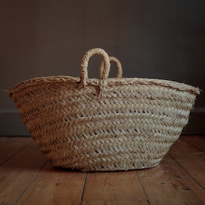 Puede incluir: Una cesta grande de mimbre natural con dos asas. La cesta está hecha de un material marrón claro y tiene una forma ligeramente redondeada.