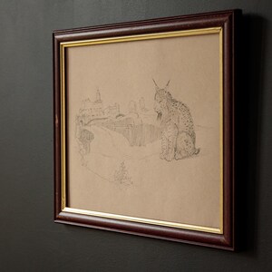 May include: Framed pencil sketch of a lynx sitting in a landscape with a building and road. The artwork is in a dark brown frame with a gold inner border. The background is a dark gray wall.