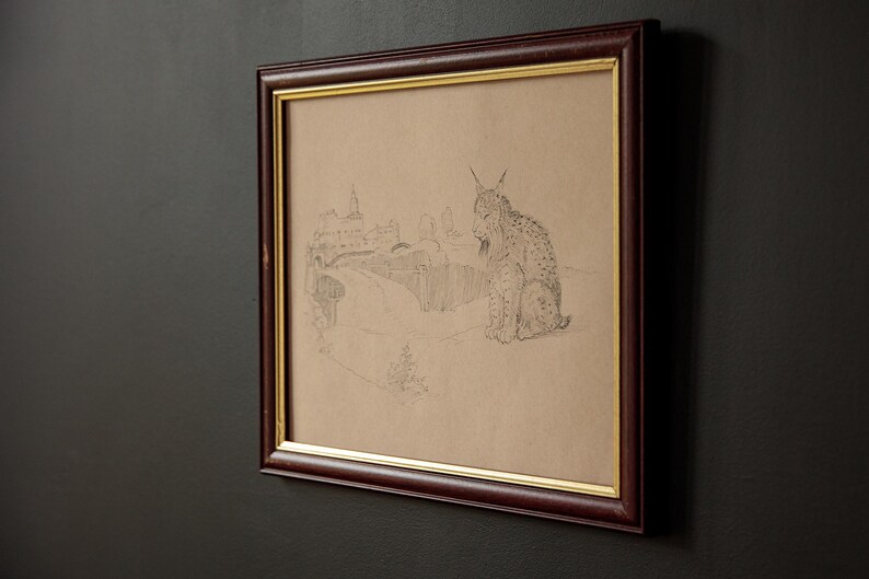 May include: Framed pencil sketch of a lynx sitting in a landscape with a building and road. The artwork is in a dark brown frame with a gold inner border. The background is a dark gray wall.