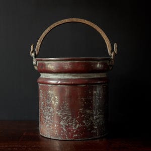 May include: An antique copper bucket with a wooden handle. The cylindrical bucket has a flared rim and a weathered, mottled finish in shades of red and brown. The arched handle is attached with metal loops. A vintage kitchenware item.