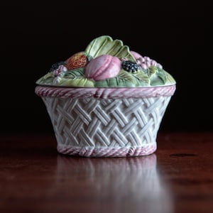 Puede incluir: Una cesta de cerámica blanca con un borde rosa, decorada con hojas verdes y frutas coloridas. La cesta tiene un patrón tejido y es perfecta para guardar objetos pequeños.