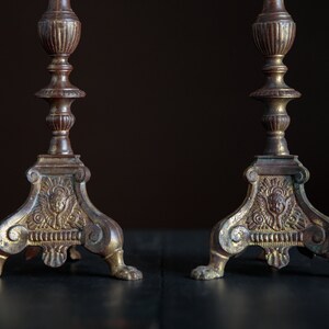 Puede incluir: Un par de candelabros antiguos de lat&oacute;n con detalles ornamentados y un acabado dorado. Cada candelabro tiene una base de tres patas con un dise&ntilde;o de querub&iacute;n.