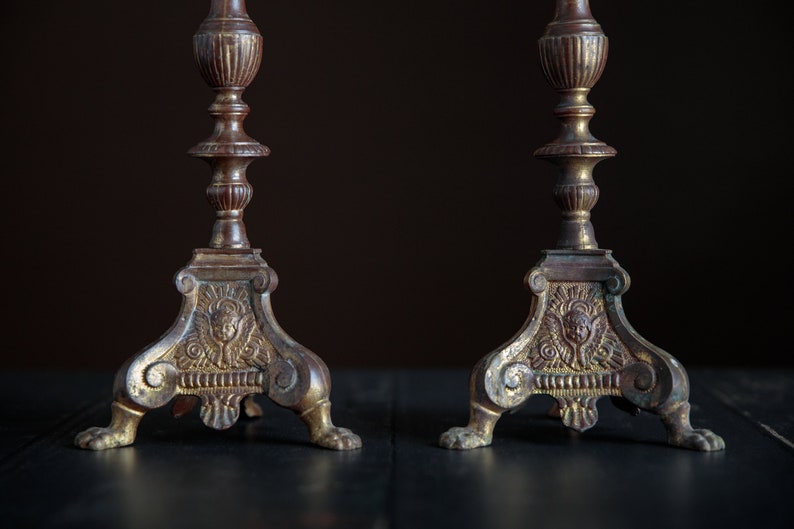 Puede incluir: Un par de candelabros antiguos de lat&oacute;n con detalles ornamentados y un acabado dorado. Cada candelabro tiene una base de tres patas con un dise&ntilde;o de querub&iacute;n.