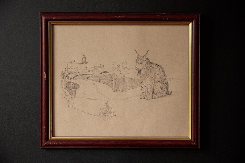 May include: Pencil sketch of a lynx sitting in front of a landscape with a building and bridge, framed in a dark red wooden frame with a gold inner border. The artwork is on tan paper.