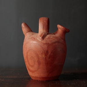 May include: A terracotta-colored ceramic jug with three spouts and a handle. The jug features a rounded body with spiral designs and is set against a dark background. The pottery appears handmade.