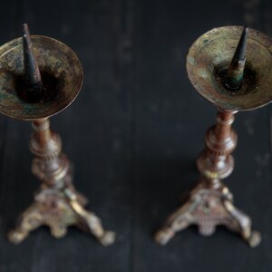 Puede incluir: Dos candelabros antiguos de lat&oacute;n con detalles ornamentados y una p&aacute;tina oscura. Cada candelabro tiene una punta puntiaguda para sujetar una vela.