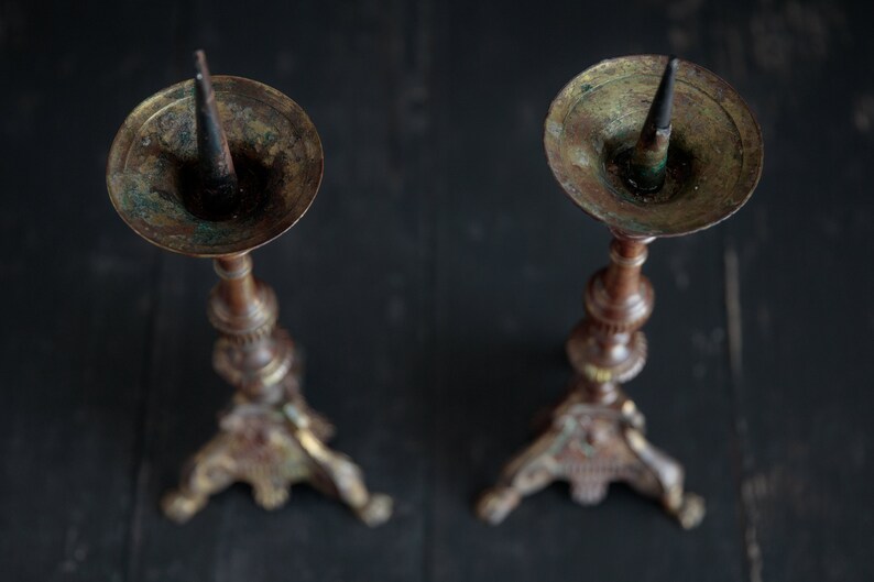 Puede incluir: Dos candelabros antiguos de lat&oacute;n con detalles ornamentados y una p&aacute;tina oscura. Cada candelabro tiene una punta puntiaguda para sujetar una vela.