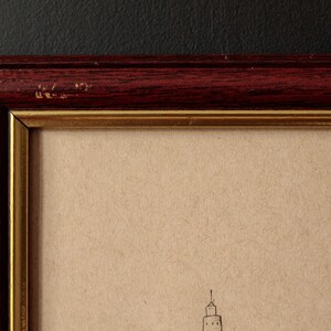 May include: Close-up of a framed artwork. The frame is a dark reddish-brown color with a gold inner border. The artwork appears to be a sketch on tan paper, featuring a building with a spire. The frame has some minor wear.