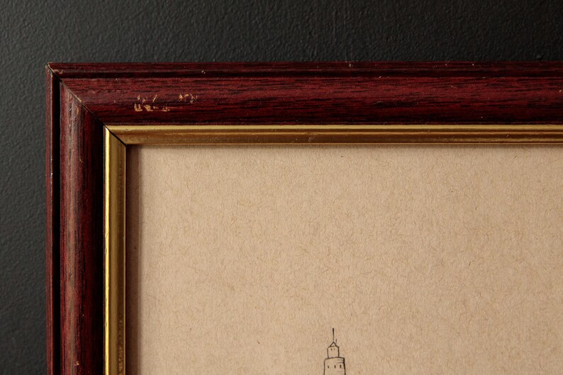 May include: Close-up of a framed artwork. The frame is a dark reddish-brown color with a gold inner border. The artwork appears to be a sketch on tan paper, featuring a building with a spire. The frame has some minor wear.