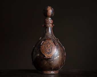 Peut inclure: Une bouteille en cuir marron avec un dessus en métal décoratif et une sangle en cuir. La bouteille a un blason avec un bouclier et un oiseau.