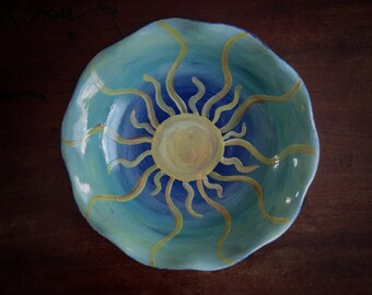 May include: A ceramic bowl with a blue and green glaze, featuring a painted sun with yellow rays. The bowl has a wavy rim.
