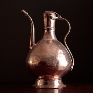May include: A copper pitcher with a hammered finish and a long spout. The pitcher has a handle that curves around the top.