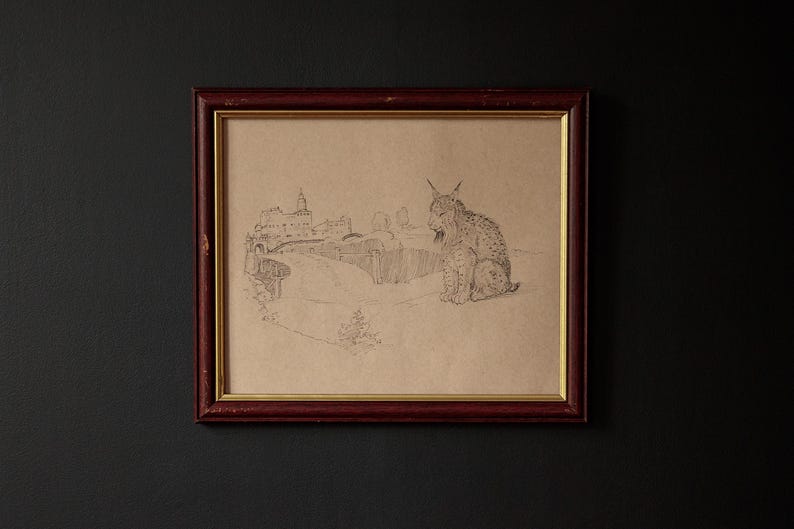 May include: A framed pencil sketch of a lynx sitting in a landscape with a building in the background. The frame is dark red with a gold inner border. The artwork is on tan paper and is mounted on a dark gray wall.