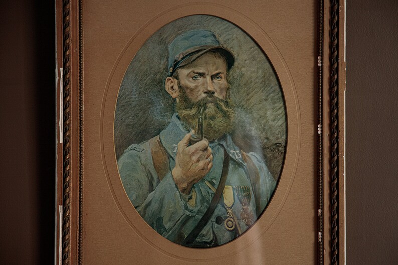 Pode incluir: Um retrato emoldurado de um homem em uniforme militar. Ele est&aacute; usando um bon&eacute; azul e tem barba. Ele est&aacute; segurando um cachimbo na m&atilde;o direita. O retrato est&aacute; em um fundo marrom claro.