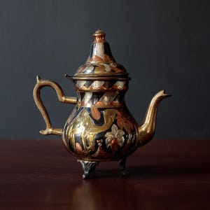 May include: An antique teapot with a lid, handle, and spout. The teapot has a gold, black, and copper colour scheme with floral and geometric patterns. It sits on three small feet and is displayed on a dark wooden surface.