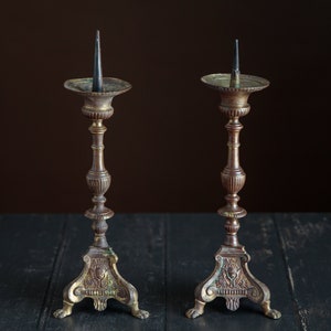 Puede incluir: Dos candelabros antiguos de lat&oacute;n con detalles ornamentados y tocones de velas negras. Los candelabros tienen una p&aacute;tina marr&oacute;n oscura y est&aacute;n sobre una superficie de madera negra.