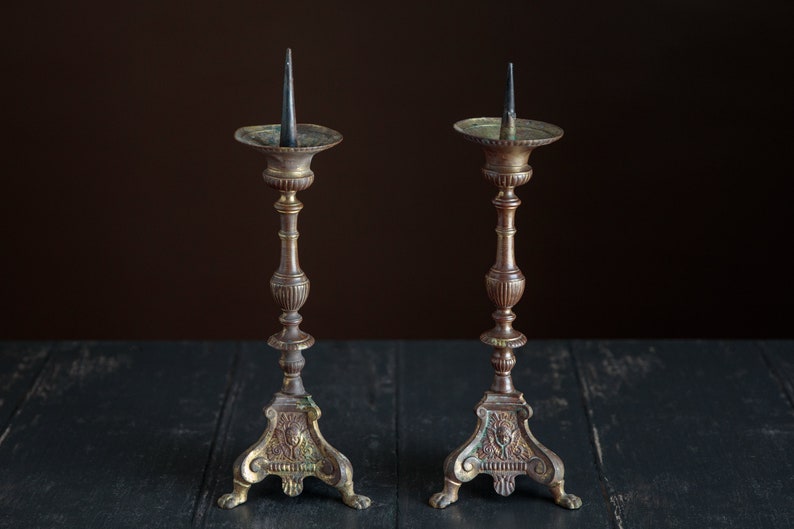 Puede incluir: Dos candelabros antiguos de lat&oacute;n con detalles ornamentados y tocones de velas negras. Los candelabros tienen una p&aacute;tina marr&oacute;n oscura y est&aacute;n sobre una superficie de madera negra.
