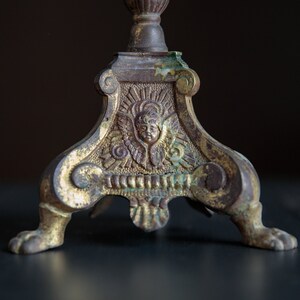 Puede incluir: Un candelero de metal dorado con un dise&ntilde;o ornamentado que presenta un &aacute;ngel con alas y una cara. El candelero tiene tres patas con pies en forma de pata.