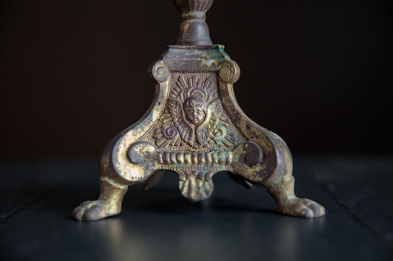 Puede incluir: Un candelero de metal dorado con un dise&ntilde;o ornamentado que presenta un &aacute;ngel con alas y una cara. El candelero tiene tres patas con pies en forma de pata.