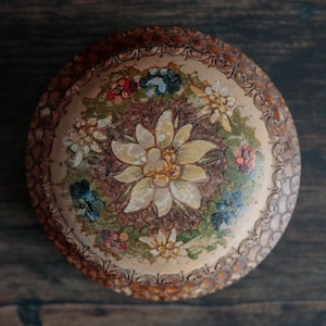 Puede incluir: Una caja redonda de madera con un diseño floral pintado en la tapa. Las flores son amarillas, blancas, rojas, azules y verdes. La caja tiene un borde de madera marrón.