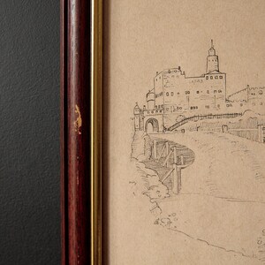 May include: A framed sketch of a castle with a tall tower, arched entrance, and bridge. The artwork is in a dark wood frame with a gold inner border. The background is a textured dark gray.