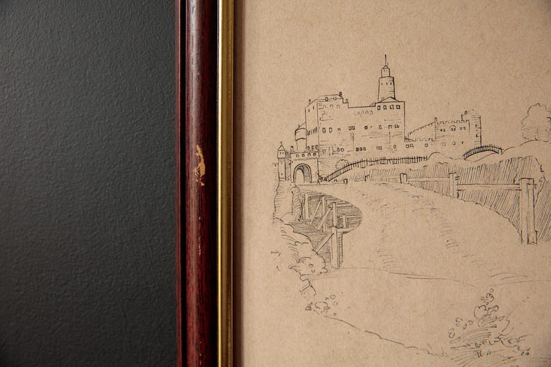 May include: A framed sketch of a castle with a tall tower, arched entrance, and bridge. The artwork is in a dark wood frame with a gold inner border. The background is a textured dark gray.