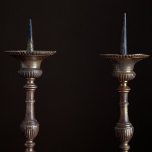 Puede incluir: Dos candelabros antiguos de lat&oacute;n con una p&aacute;tina marr&oacute;n oscura. Cada candelero tiene un tallo alto y delgado y una base circular ancha. Los candelabros est&aacute;n vac&iacute;os y tienen una punta puntiaguda en la parte superior.
