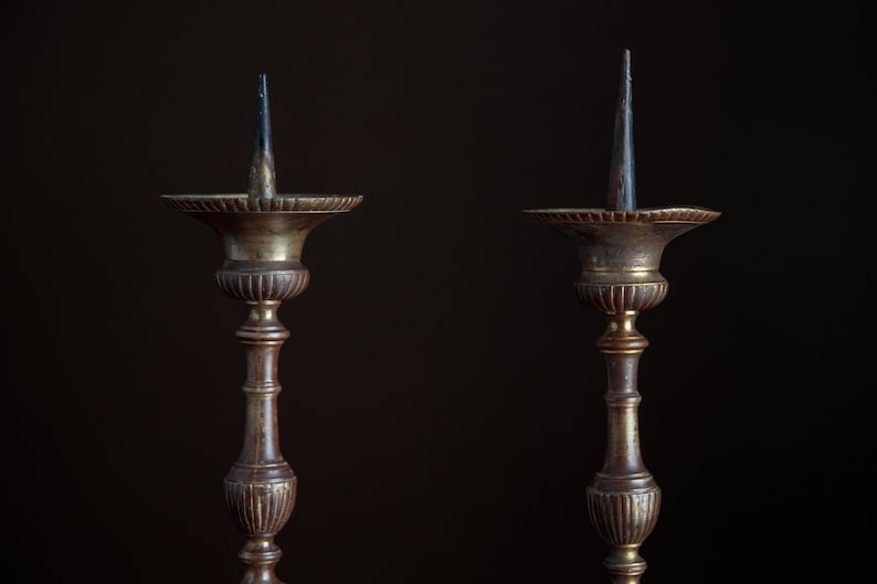 Puede incluir: Dos candelabros antiguos de lat&oacute;n con una p&aacute;tina marr&oacute;n oscura. Cada candelero tiene un tallo alto y delgado y una base circular ancha. Los candelabros est&aacute;n vac&iacute;os y tienen una punta puntiaguda en la parte superior.