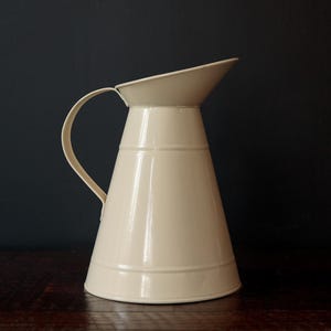 Puede incluir: Jarra de metal color crema con asa curva y boquilla acampanada. La jarra tiene forma cónica con bandas horizontales alrededor del cuerpo. El fondo es de color oscuro y sólido, resaltando la forma de la jarra.