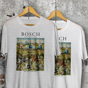 May include: Two t-shirts with a print of Hieronymus Bosch's "The Garden of Earthly Delights" painting. The left t-shirt is grey and the right t-shirt is white. Both t-shirts have the word "BOSCH" printed above the painting.