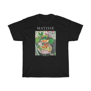 May include: Black t-shirt with a print of a painting by Henri Matisse titled "Goldfish and a Bowl of Flowers". The painting depicts a bowl of goldfish on a table with flowers and leaves.
