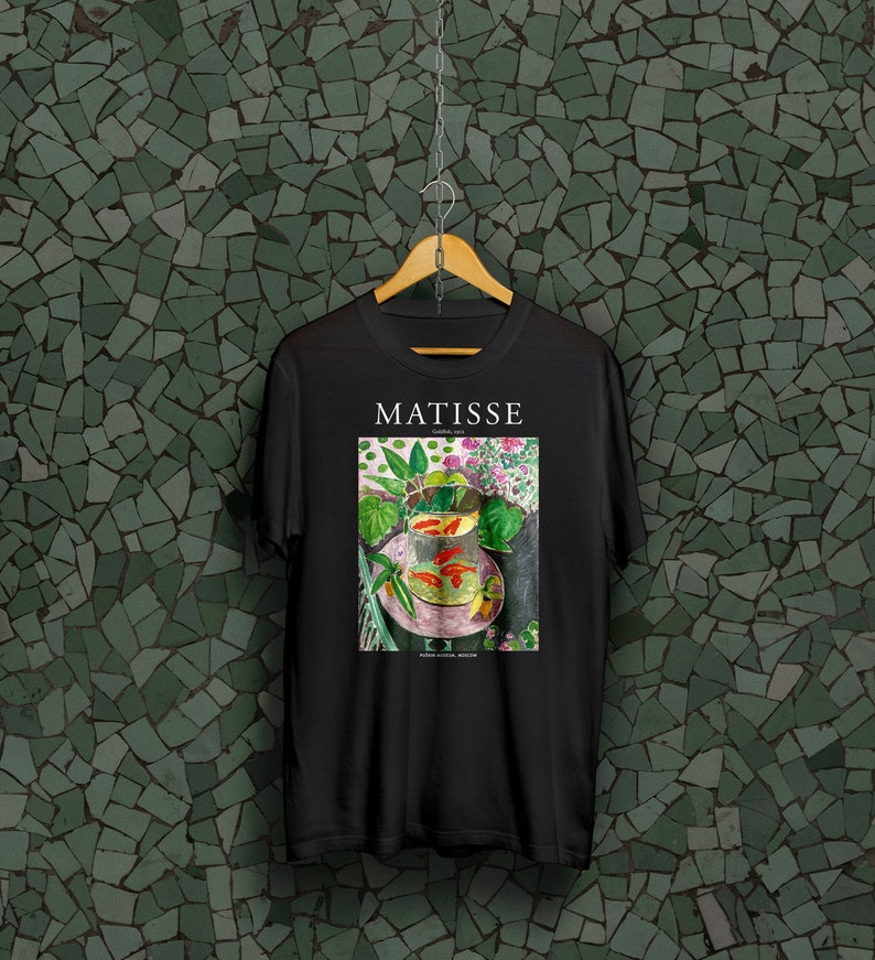 May include: Black t-shirt with a graphic print of a painting by Henri Matisse titled "Goldfish and Sculpture". The painting depicts a bowl of goldfish with green plants and a sculpture in the background. The text "MATISSE" and "Goldfish 1912" is printed above the painting.