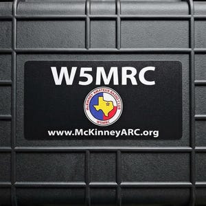May include: A black hard-shell protective equipment case with a grid-patterned exterior and a top handle. A rectangular sticker features the white text W5MRC, a circular McKinney Amateur Radio Club logo with a Texas map, and the URL www.McKinneyARC.org.