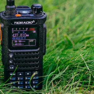 May include: A black TIDRADIO handheld radio with an orange accent is displayed on a bed of green grass. The radio's screen shows numbers and text, including "437.810bb" and "CH-012 S". The radio has buttons and a keypad.