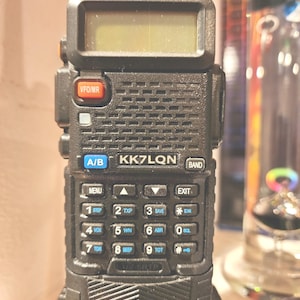 May include: A black handheld radio with a digital display and numerous buttons. The radio has the text "KK7LQN" and various function labels like "MENU", "EXIT", and "SCAN". It is designed for communication and likely used for amateur radio or similar purposes.