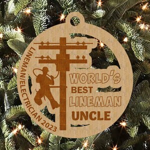 May include: A brown wooden Christmas ornament with a silhouette of a lineman working on a power pole. The ornament reads "World's Best Lineman Uncle" and "Lineman/Electrician 2023".
