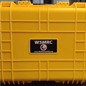 May include: A bright yellow, rectangular, hard-shell protective equipment case with a grid-like reinforced exterior. A black rectangular label is centered on the front, displaying the white text W5MRC, a circular club logo, and the URL www.McKinneyARC.org.