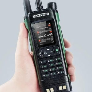 May include: A black and green Baofeng two-way radio held in a hand. The radio has a screen displaying channel information and a keypad with numbers and function keys. The radio's antenna is visible.