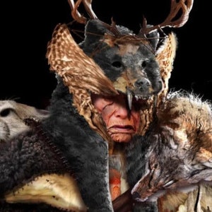 May include: A person wearing a costume made of animal furs and hides. The costume includes a bear head, deer antlers, and a wolf pelt. The person's face is painted with red and white markings.