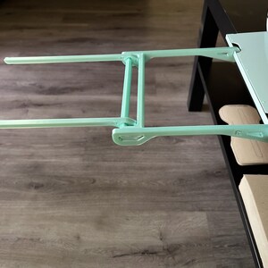 May include: A light blue metal extension arm with two horizontal bars. The arm is attached to a white surface and is extended out over a wooden floor.