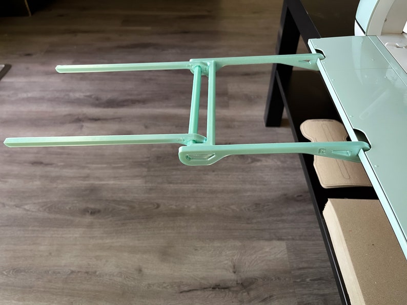 May include: A light blue metal extension arm with two horizontal bars. The arm is attached to a white surface and is extended out over a wooden floor.