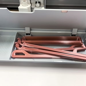 May include: A close-up of a gray drawer with a rose gold colored plastic tool inside. The tool is a long, thin, rectangular shape with a hook on one end.